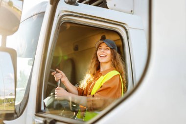 Yansıtıcı yelek giyen genç bir kadın sürücü büyük bir kamyonun direksiyonunda oturuyor. Şapkalı bir kadın büyük bir arabanın sürücü koltuğunda oturur. Ulaşım, iş kavramı.