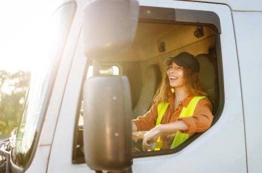 Yansıtıcı yelek giyen genç bir kadın sürücü büyük bir kamyonun direksiyonunda oturuyor. Şapkalı bir kadın büyük bir arabanın sürücü koltuğunda oturur. Ulaşım, iş kavramı.