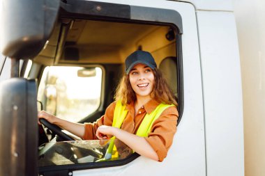 Yansıtıcı yelek giyen genç bir kadın sürücü büyük bir kamyonun direksiyonunda oturuyor. Şapkalı bir kadın büyük bir arabanın sürücü koltuğunda oturur. Ulaşım, iş kavramı.
