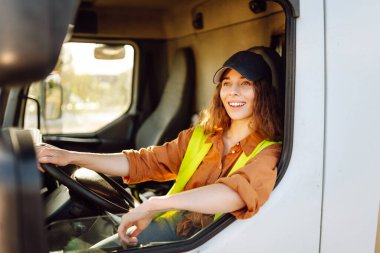 Yansıtıcı yelek giyen genç bir kadın sürücü büyük bir kamyonun direksiyonunda oturuyor. Şapkalı bir kadın büyük bir arabanın sürücü koltuğunda oturur. Ulaşım, iş kavramı.