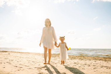 Mutlu anne ve kızı güneşli bir sahilde birlikte geziyorlar. Güzel bir kadın ve çocuğu deniz manzaralı sahilde gün batımının tadını çıkarıyorlar. Aile, eğlence ve çocukluk kavramı.