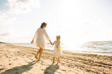 Mutlu anne ve kızı güneşli bir sahilde birlikte geziyorlar. Güzel bir kadın ve çocuğu deniz manzaralı sahilde gün batımının tadını çıkarıyorlar. Aile, eğlence ve çocukluk kavramı.