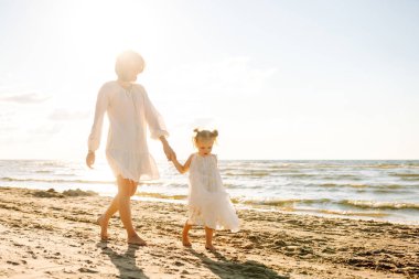 Mutlu anne ve kızı güneşli bir sahilde birlikte geziyorlar. Güzel bir kadın ve çocuğu deniz manzaralı sahilde gün batımının tadını çıkarıyorlar. Aile, eğlence ve çocukluk kavramı.