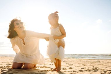 Mutlu anne ve kızı güneşli bir sahilde birlikte geziyorlar. Güzel bir kadın ve çocuğu deniz manzaralı sahilde gün batımının tadını çıkarıyorlar. Aile, eğlence ve çocukluk kavramı.