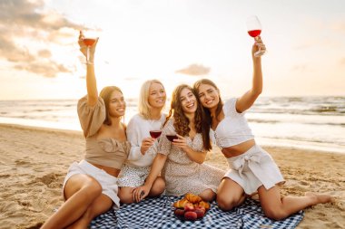 Neşeli bayan arkadaşlar gün batımı plajında piknikte birlikte vakit geçiriyorlar. Ellerinde gözlüklerle deniz manzarasının tadını çıkaran dört genç kadın. Arkadaşlık ve eğlence konsepti.