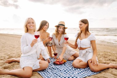 Neşeli bayan arkadaşlar gün batımı plajında piknikte birlikte vakit geçiriyorlar. Ellerinde gözlüklerle deniz manzarasının tadını çıkaran dört genç kadın. Arkadaşlık ve eğlence konsepti.