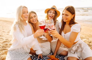 Neşeli bayan arkadaşlar gün batımı plajında piknikte birlikte vakit geçiriyorlar. Ellerinde gözlüklerle deniz manzarasının tadını çıkaran dört genç kadın. Arkadaşlık ve eğlence konsepti.