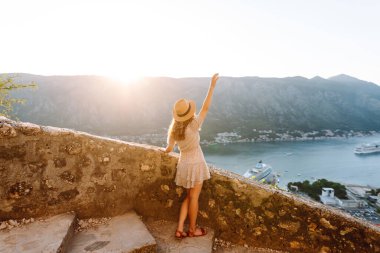 Deniz koyunun manzarasının keyfini çıkaran mutlu bir kadının dikiz görüntüsü. Şapkalı bir gezgin kendini özgür hisseder ve bir dağın tepesinde oturur. Turizm, seyahat konsepti. Etkin yaşam biçimi.