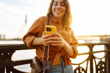 Gün batımında genç bir kadın elinde telefonla setin kenarında yürüyor. Güzel bir kadın blog yazıyor ve açık havanın tadını çıkarıyor. Teknoloji ve yürüme kavramı.