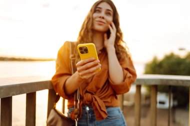 Gün batımında genç bir kadın elinde telefonla setin kenarında yürüyor. Güzel bir kadın blog yazıyor ve açık havanın tadını çıkarıyor. Teknoloji ve yürüme kavramı.