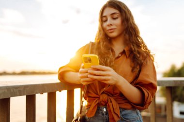 Gün batımında genç bir kadın elinde telefonla setin kenarında yürüyor. Güzel bir kadın blog yazıyor ve açık havanın tadını çıkarıyor. Teknoloji ve yürüme kavramı.