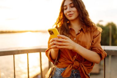 Gün batımında genç bir kadın elinde telefonla setin kenarında yürüyor. Güzel bir kadın blog yazıyor ve açık havanın tadını çıkarıyor. Teknoloji ve yürüme kavramı.