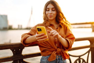 Gün batımında genç bir kadın elinde telefonla setin kenarında yürüyor. Güzel bir kadın blog yazıyor ve açık havanın tadını çıkarıyor. Teknoloji ve yürüme kavramı.