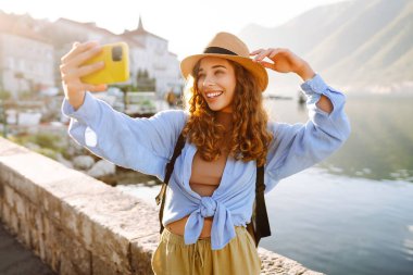 Şapkalı, şapkalı genç, kıvırcık bir kadın arka planda deniz kenarı olan bir selfie çekiyor. Sırt çantası ve şapkası olan bir bayan gezgin gün doğumunda deniz manzarasının tadını çıkarıyor. Macera konsepti
