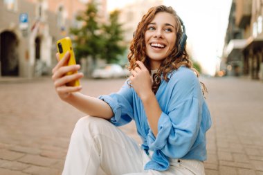 Kıvırcık saçlı, kulaklık takan ve cep telefonu kullanan güzel bir kadın şehir caddesinde eğleniyor. Genç bir kadın müzik dinlemekten ve dışarıda gün batımını izlemekten hoşlanır. Turizm konsepti.
