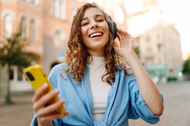 Kıvırcık saçlı, kulaklık takan ve cep telefonu kullanan güzel bir kadın şehir caddesinde eğleniyor. Genç bir kadın müzik dinlemekten ve dışarıda gün batımını izlemekten hoşlanır. Turizm konsepti.