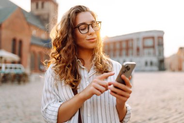 Gün batımı caddesinde elinde telefon tutan gözlüklü güzel bir kadının portresi. Güzel bir kadın telefonda mesajlaşıyor, blog yazıyor ve şehirde yürüyüş yapmaktan zevk alıyor. Turizm konsepti.