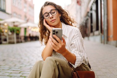 Gün batımı caddesinde elinde telefon tutan gözlüklü güzel bir kadının portresi. Güzel bir kadın telefonda mesajlaşıyor, blog yazıyor ve şehirde yürüyüş yapmaktan zevk alıyor. Turizm konsepti.