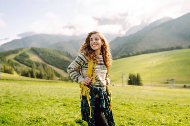 Sırtında sarı sırt çantasıyla yeşil çayırda duran güzel bayan gezgin. Genç bir kadın gün batımının tadını çıkarıyor. Keyif ve rahatlama konsepti.