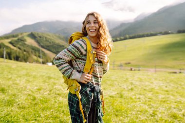 Sırtında sarı sırt çantasıyla yeşil çayırda duran güzel bayan gezgin. Genç bir kadın gün batımının tadını çıkarıyor. Keyif ve rahatlama konsepti.