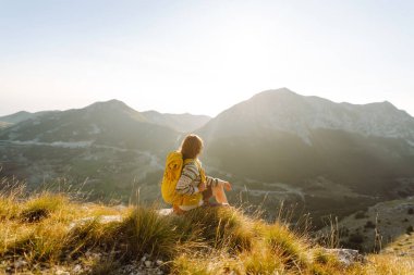 Renkli bir sırt çantasıyla dağ zirvesi boyunca yürüyen genç bir bayan gezgin. Neşeli bir kadın kendini özgür hisseder ve gün batımında dağ manzarasının tadını çıkarır. Yürüyüş konsepti. Etkin yaşam biçimi.