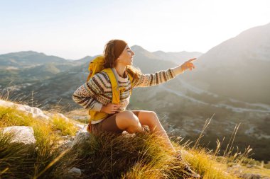 Renkli bir sırt çantasıyla dağ zirvesi boyunca yürüyen genç bir bayan gezgin. Neşeli bir kadın kendini özgür hisseder ve gün batımında dağ manzarasının tadını çıkarır. Yürüyüş konsepti. Etkin yaşam biçimi.