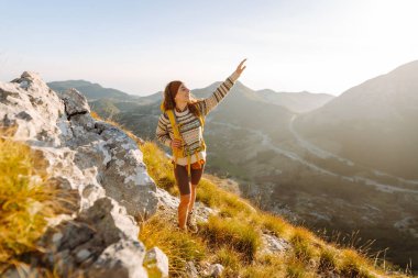 Renkli bir sırt çantasıyla dağ zirvesi boyunca yürüyen genç bir bayan gezgin. Neşeli bir kadın kendini özgür hisseder ve gün batımında dağ manzarasının tadını çıkarır. Yürüyüş konsepti. Etkin yaşam biçimi.