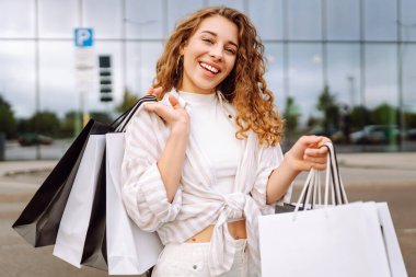 Alışveriş torbalarıyla modern bir alışveriş merkezinin arka planında duran mutlu kadın. Alışveriş zamanı! Alışverişten ve açık havadan hoşlanan güzel bir kadın. Tüketim kavramı.