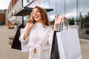 Alışveriş torbalarıyla modern bir alışveriş merkezinin arka planında duran mutlu kadın. Alışveriş zamanı! Alışverişten ve açık havadan hoşlanan güzel bir kadın. Tüketim kavramı.