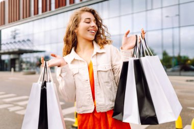 Alışveriş torbalarıyla modern bir alışveriş merkezinin arka planında duran mutlu kadın. Alışveriş zamanı! Alışverişten ve açık havadan hoşlanan güzel bir kadın. Tüketim kavramı.