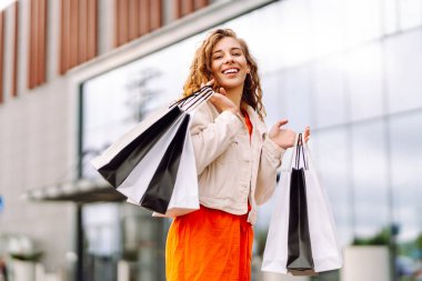 Alışveriş torbalarıyla modern bir alışveriş merkezinin arka planında duran mutlu kadın. Alışveriş zamanı! Alışverişten ve açık havadan hoşlanan güzel bir kadın. Tüketim kavramı.
