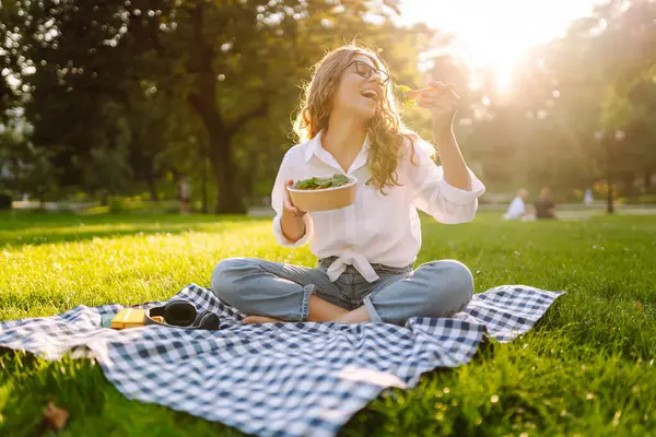 Gözlüklü mutlu kadın piknik paspasında salata yiyor. Sağlıklı bir kasenin tadını çıkaran ve güneşli bir parkta güneşlenen genç bir kadın. Rahatlama ve keyif kavramı.
