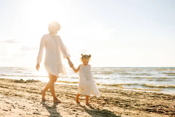 Mutlu anne ve kızı güneşli bir sahilde birlikte geziyorlar. Güzel bir kadın ve çocuğu deniz manzaralı sahilde gün batımının tadını çıkarıyorlar. Aile, eğlence ve çocukluk kavramı.
