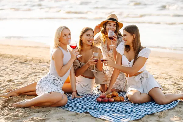 Neşeli bayan arkadaşlar gün batımı plajında piknikte birlikte vakit geçiriyorlar. Ellerinde gözlüklerle deniz manzarasının tadını çıkaran dört genç kadın. Arkadaşlık ve eğlence konsepti.