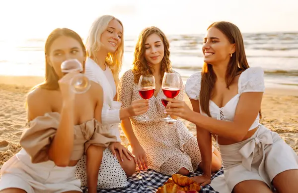 Neşeli bayan arkadaşlar gün batımı plajında piknikte birlikte vakit geçiriyorlar. Ellerinde gözlüklerle deniz manzarasının tadını çıkaran dört genç kadın. Arkadaşlık ve eğlence konsepti.