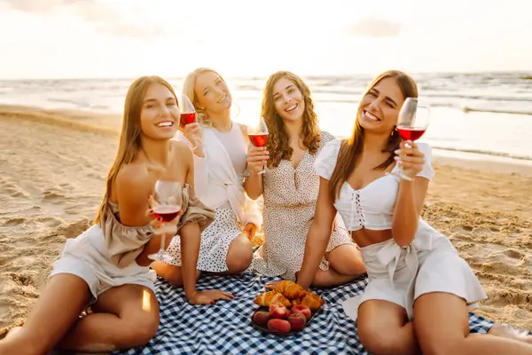Neşeli bayan arkadaşlar gün batımı plajında piknikte birlikte vakit geçiriyorlar. Ellerinde gözlüklerle deniz manzarasının tadını çıkaran dört genç kadın. Arkadaşlık ve eğlence konsepti.