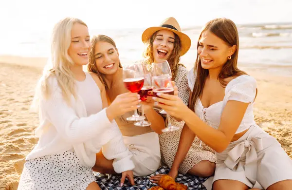 Neşeli bayan arkadaşlar gün batımı plajında piknikte birlikte vakit geçiriyorlar. Ellerinde gözlüklerle deniz manzarasının tadını çıkaran dört genç kadın. Arkadaşlık ve eğlence konsepti.