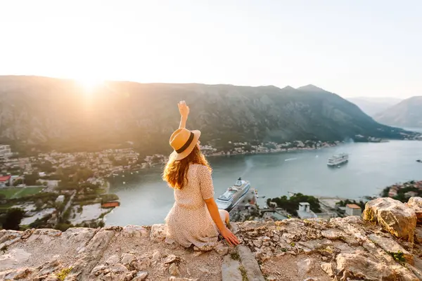 Deniz koyunun manzarasının keyfini çıkaran mutlu bir kadının dikiz görüntüsü. Şapkalı bir gezgin kendini özgür hisseder ve bir dağın tepesinde oturur. Turizm, seyahat konsepti. Etkin yaşam biçimi.