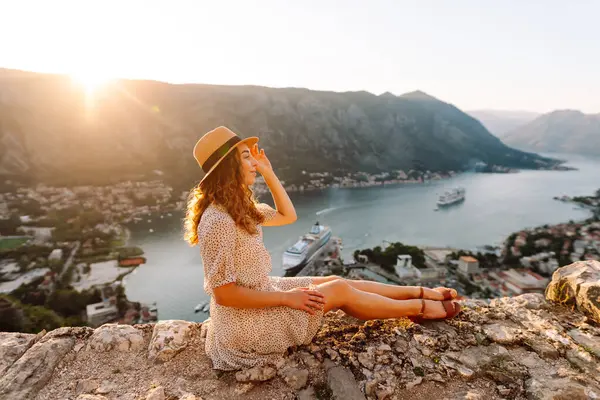 Deniz koyunun manzarasının keyfini çıkaran mutlu bir kadının dikiz görüntüsü. Şapkalı bir gezgin kendini özgür hisseder ve bir dağın tepesinde oturur. Turizm, seyahat konsepti. Etkin yaşam biçimi.