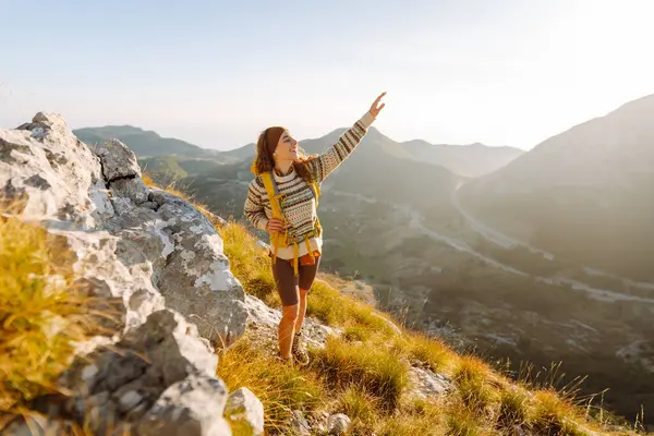 Renkli bir sırt çantasıyla dağ zirvesi boyunca yürüyen genç bir bayan gezgin. Neşeli bir kadın kendini özgür hisseder ve gün batımında dağ manzarasının tadını çıkarır. Yürüyüş konsepti. Etkin yaşam biçimi.