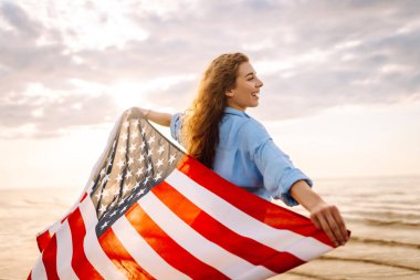 Kumsalda omuzlarında Amerikan bayrağı olan mutlu bir kadın. Denizde gün batımının keyfini çıkaran güzel bir kadın, özgürlük bayrağını taşıyor. Bağımsızlık ve kutlama kavramı.