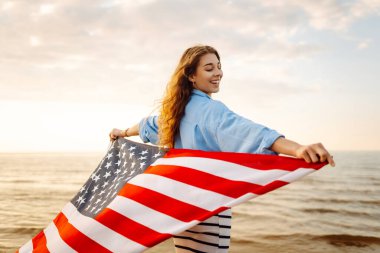 Kumsalda omuzlarında Amerikan bayrağı olan mutlu bir kadın. Denizde gün batımının keyfini çıkaran güzel bir kadın, özgürlük bayrağını taşıyor. Bağımsızlık ve kutlama kavramı.