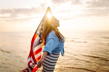 Kumsalda omuzlarında Amerikan bayrağı olan mutlu bir kadın. Denizde gün batımının keyfini çıkaran güzel bir kadın, özgürlük bayrağını taşıyor. Bağımsızlık ve kutlama kavramı.