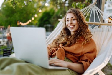 Güneşli bir parkta hamakta dizüstü bilgisayarlı gülümseyen bir kadın. Hamakta çalışan serbest çalışan güzel bir bayan. Teknoloji kavramı, uzaktan çalışma, rahatlama..