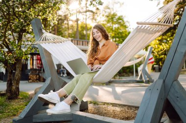 Güneşli bir parkta hamakta dizüstü bilgisayarlı gülümseyen bir kadın. Hamakta çalışan serbest çalışan güzel bir bayan. Teknoloji kavramı, uzaktan çalışma, rahatlama..