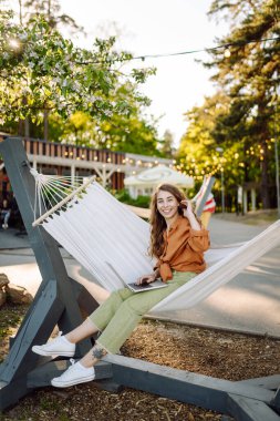 Güneşli bir parkta hamakta dizüstü bilgisayarlı gülümseyen bir kadın. Hamakta çalışan serbest çalışan güzel bir bayan. Teknoloji kavramı, uzaktan çalışma, rahatlama..