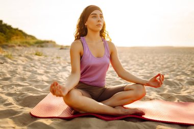 Spor kıyafetli genç bir kadın pembe yoga minderinde esneme egzersizleri yapıyor. Rahat kadın yoga yapıyor ve gün batımında deniz manzarasının tadını çıkarıyor. Rahatlama kavramı.