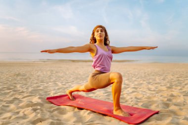 Spor kıyafetli genç bir kadın pembe yoga minderinde esneme egzersizleri yapıyor. Rahat kadın yoga yapıyor ve gün batımında deniz manzarasının tadını çıkarıyor. Rahatlama kavramı.