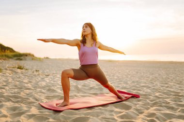 Spor kıyafetli genç bir kadın pembe yoga minderinde esneme egzersizleri yapıyor. Rahat kadın yoga yapıyor ve gün batımında deniz manzarasının tadını çıkarıyor. Rahatlama kavramı.