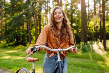 Ormanda, güneşin altında bisiklet süren mutlu bir kadın var. Güzel bir kadın gün batımında bisiklet sürmekten zevk alıyor, dinleniyor. Aktif yaşam tarzı. Hafta sonu konsepti.